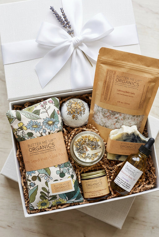 A curated self-care gift set from Butter Me Up Organics arranged in a white gift box with a large white ribbon. The set includes a bath bomb, a bath salt pouch, a bath towel, a candle, a small jar, a dropper bottle, and a soap bar, all in natural kraft paper packaging with botanical illustrations.